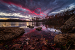 A serene lake reflecting a dramatic sunset with pink and purple clouds, surrounded by autumn trees and rocks.