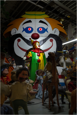 A colorful clown stands in front of a large clown backdrop, surrounded by various puppets and figures.