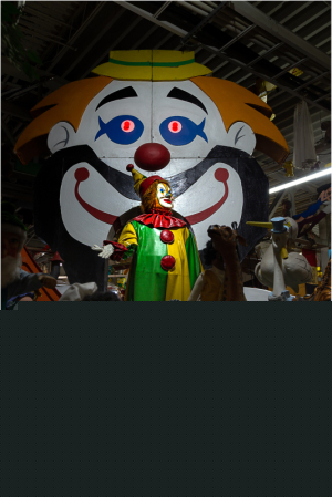 A colorful clown stands in front of a large clown backdrop, surrounded by various puppets and figures.