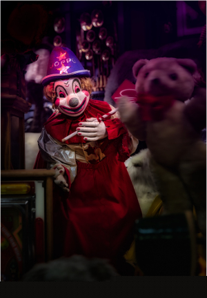 A creepy clown in a red dress, holding a toy, surrounded by shadows.