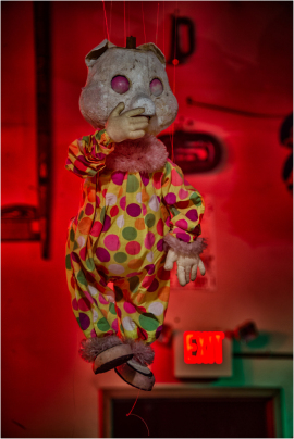A haunting puppet dressed in a polka-dotted clown suit, suspended in eerie red light.