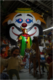 A colorful clown stands in front of a large clown backdrop, surrounded by various puppets and figures.