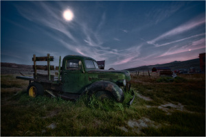 Abandoned green truck overgrown with grass under a twilight sky.