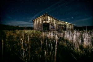 A weathered barn stands under a starry sky, surrounded by tall grass.