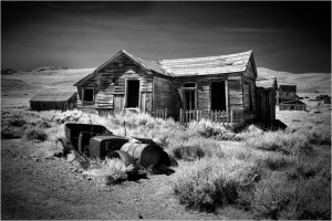 Abandoned wooden house in a ghost town, surrounded by overgrown grass and rusted barrels.
