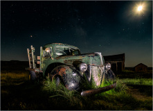 An old, rusted truck sits in a field under a starry night sky.