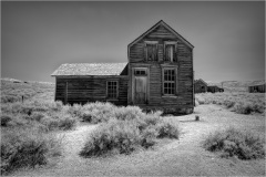 Abandoned wooden house in a ghost town, surrounded by dry grass and distant structures.