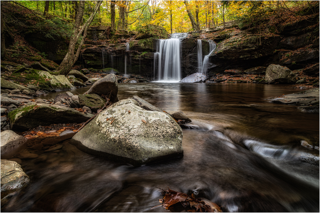 A serene waterfall cascades over rocks into a tranquil pool, surrounded by autumn foliage.