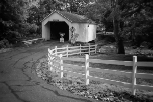 A black and white image of a covered bridge with a winding road leading to it.
