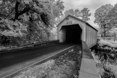 A black and white image of a covered bridge surrounded by trees.