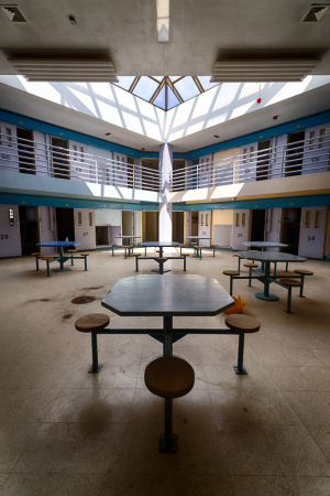Empty prison cafeteria with tables and chairs, harsh lighting, no people around.