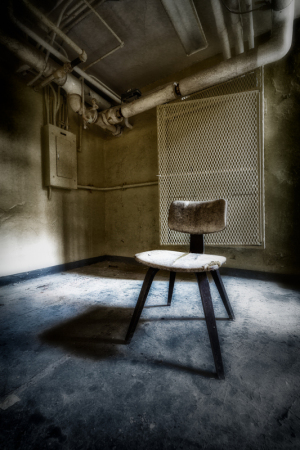 A solitary, worn-out wooden chair stands in a shadowy corner of an eerie room.