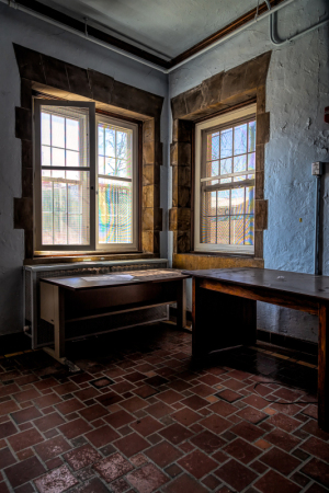 A corner room with two windows, wooden tables, and brick flooring; a blend of old charm and neglect.