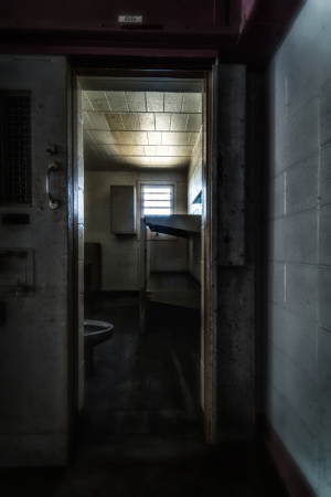 A dimly lit prison cell with a narrow doorway and a single bed.