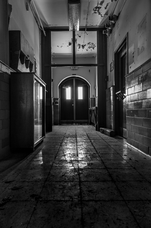 A dimly lit hallway with peeling walls and a shiny floor leading to double doors.