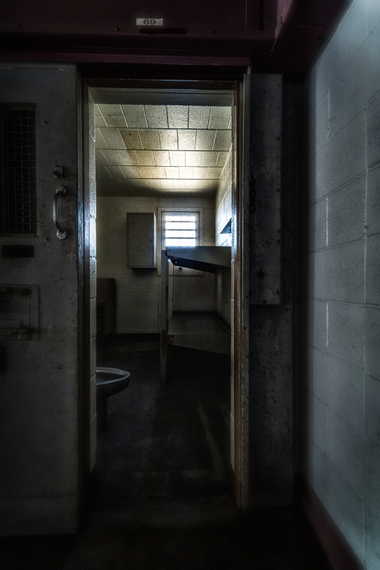 A dimly lit prison cell with a narrow doorway and a single bed.