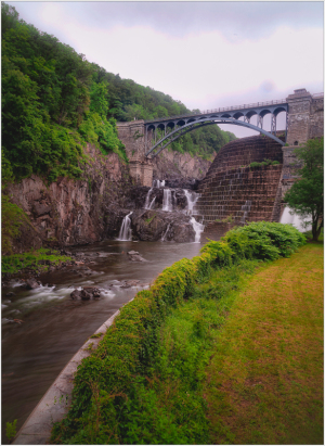 A waterfall cascades down rocky cliffs under a bridge surrounded by lush greenery.