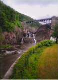 A waterfall cascades down rocky cliffs under a bridge surrounded by lush greenery.