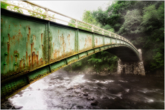 Rusty green bridge arching over a misty river, surrounded by lush trees.