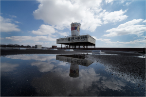 A tall water tower stands on a desolate rooftop, casting a long shadow over puddles.