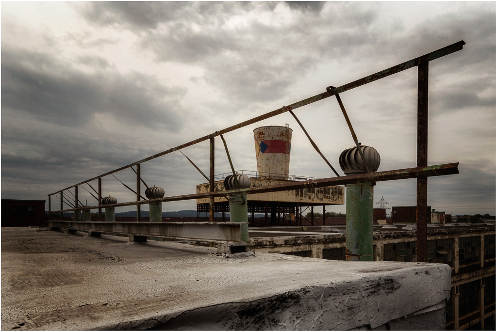 A rooftop with a rusty railing and a distant water tower under a cloudy sky.