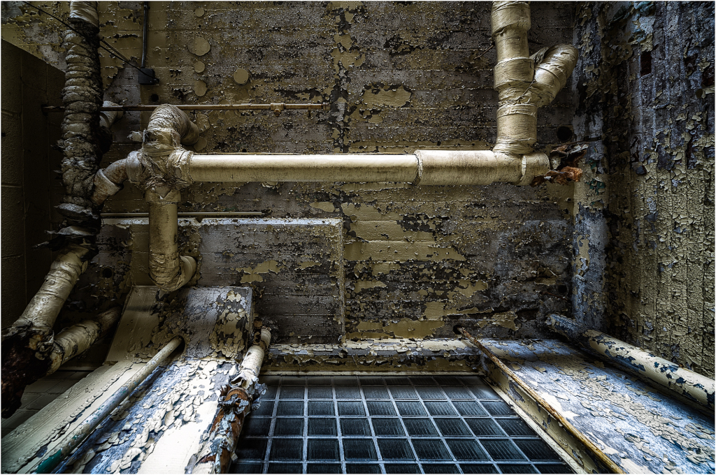 A gritty, peeling wall with old pipes and a dark grid pattern beneath.