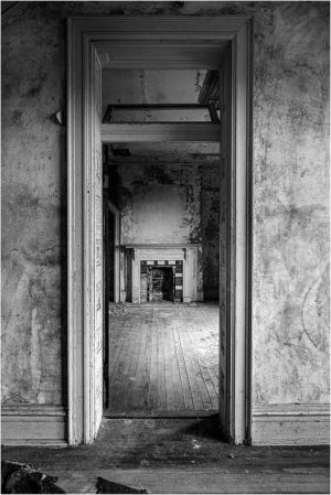 Black and white photo of an empty room through a doorway, showing peeling walls and a fireplace.