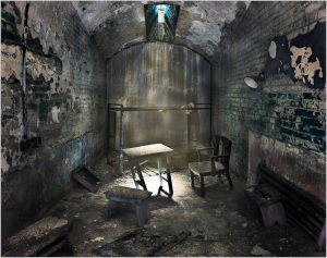 A dimly lit, abandoned cell with a table and chairs in disrepair.