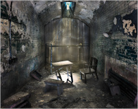 A dimly lit, abandoned cell with a table and chairs in disrepair.