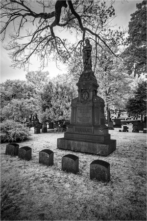 A weathered gravestone stands in a quiet cemetery, surrounded by shadowy trees.