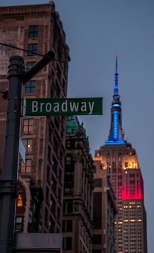 Broadway street sign with Empire State Building illuminated in red, white, and blue at dusk.