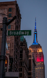 Broadway street sign with Empire State Building illuminated in red, white, and blue at dusk.