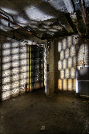 An empty, dimly lit room with peeling walls and shadows cast by barred windows.