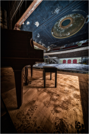 A grand piano and a small stool on a dusty theater stage with ornate ceiling details.