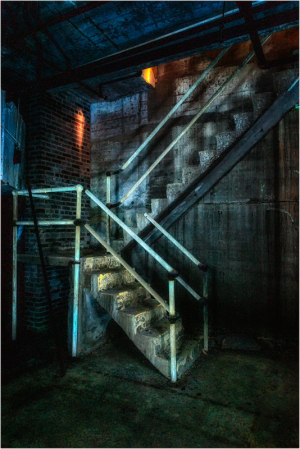 A dimly lit, worn staircase in an industrial setting with peeling paint and shadows.