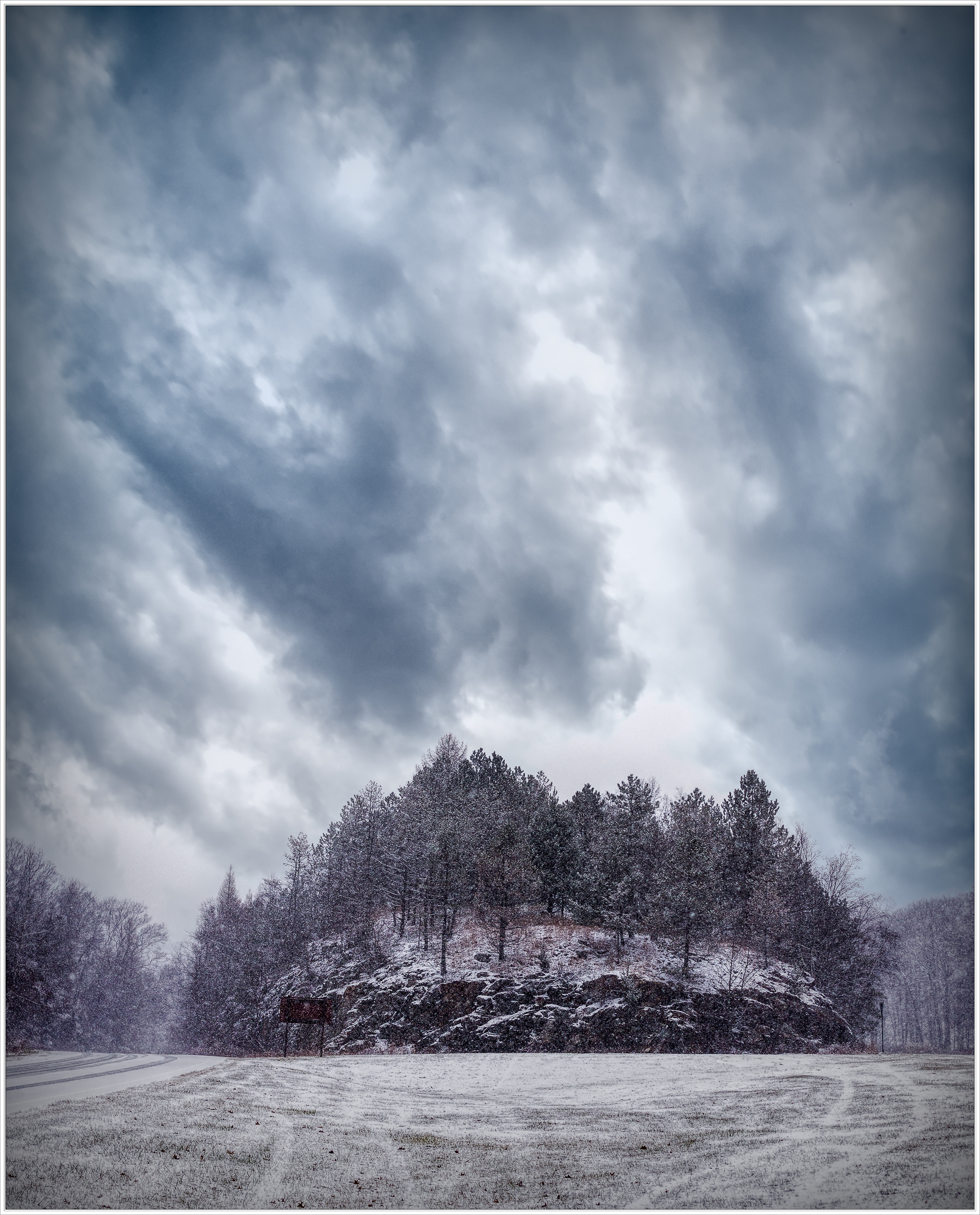 A snow-covered hill looms under dramatic clouds, trees dressed in frost, a chilling stillness prevails.