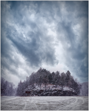A snow-covered hill looms under dramatic clouds, trees dressed in frost, a chilling stillness prevails.