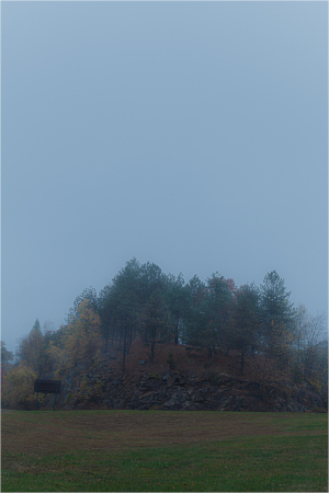 A fog-shrouded mound amidst desolate fields, crowned with spectral trees.