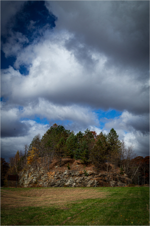 A forbidding rocky hill crowned with trees under ominous clouds.