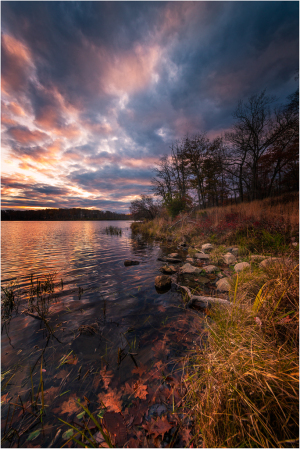 A tranquil lakeside at dusk, with vibrant clouds reflecting on the water's surface.