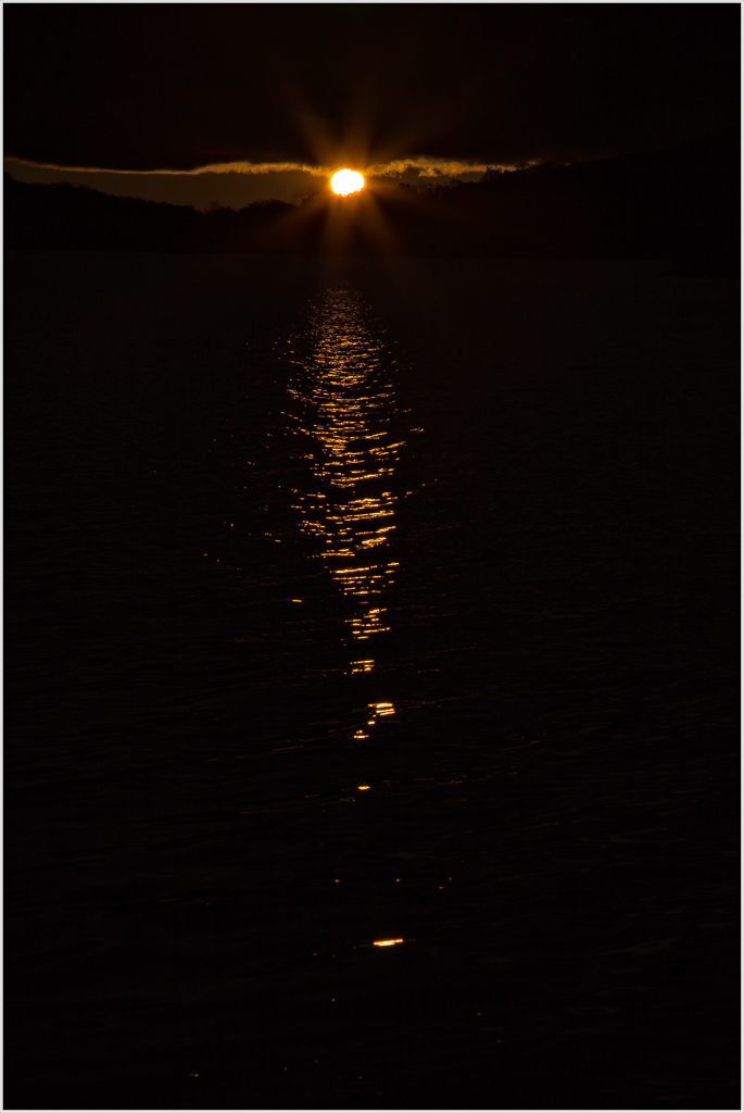 A glowing sun rises above dark waters, casting shimmering reflections.