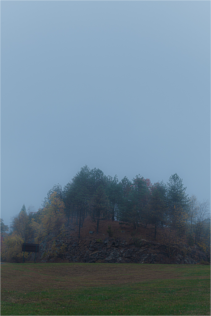 A fog-shrouded mound amidst desolate fields, crowned with spectral trees.