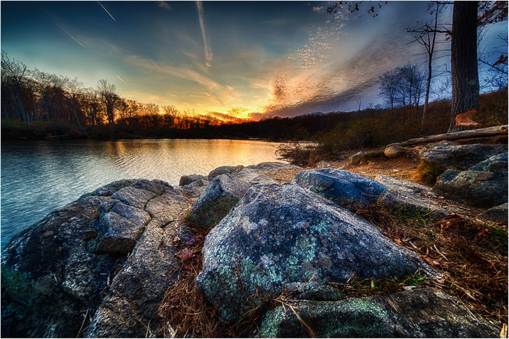 A tranquil lakeside scene at sunset with rocks and trees reflecting warm colors.