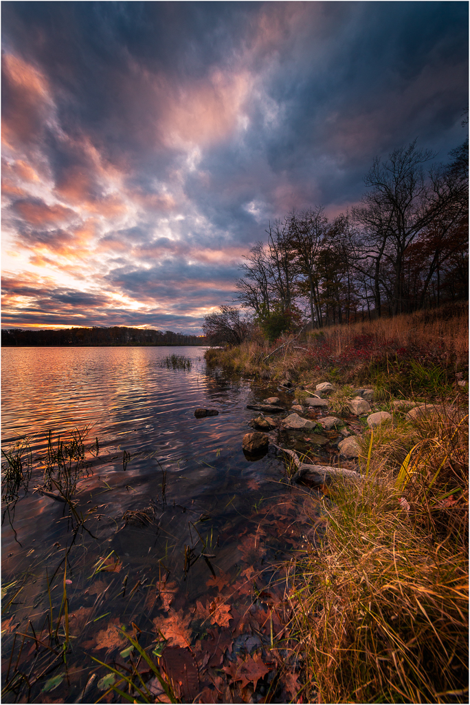 A tranquil lakeside at dusk, with vibrant clouds reflecting on the water's surface.