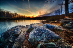 A tranquil lakeside scene at sunset with rocks and trees reflecting warm colors.