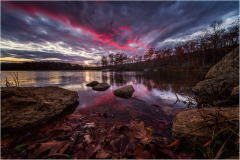A tranquil lake reflects a vibrant sunset amidst rocky shores and autumn leaves.