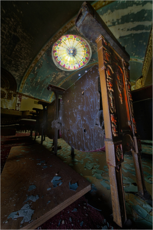 An ornate wooden table stands askew beneath a stained glass window, dust and debris scattered around.