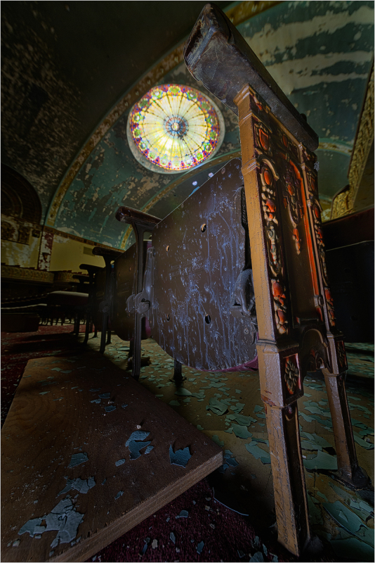 An ornate wooden table stands askew beneath a stained glass window, dust and debris scattered around.