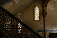 Stained glass window illuminates a worn staircase with ornate iron railing.