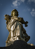A weathered statue of a figure with arms raised, holding a cross against a clear sky.